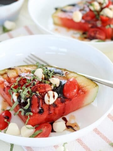 A white plate of grilled watermelon caprese wedge drizzled with hot honey.