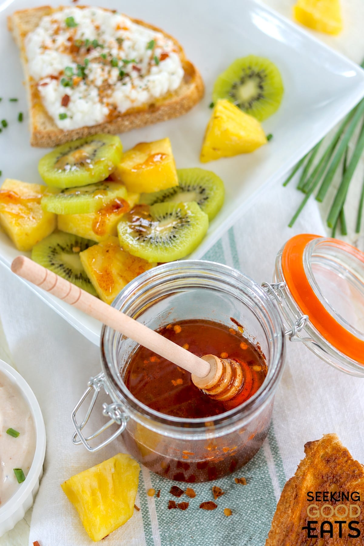 Hot honey in a clear jar by a plate of whipped cottage cheese toast and mixed fruit drizzled with hot honey.