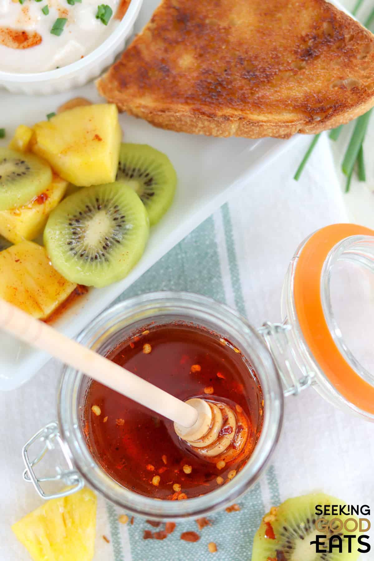 A jar of hot honey with a honey spoon by a white plate of fruit and toast.