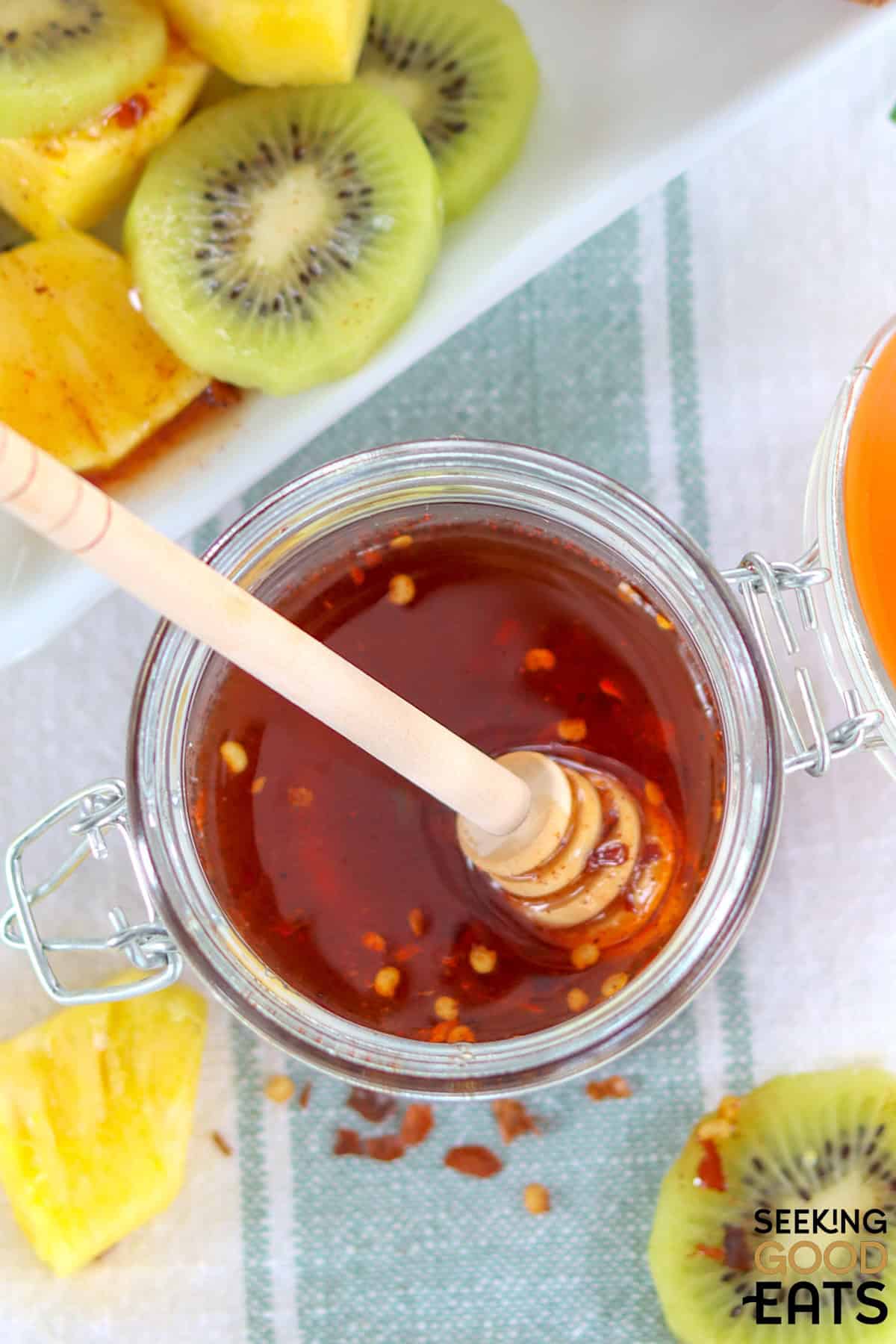 A jar of hot honey next to kiwi and pineapple drizzled with hot honey.