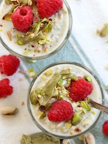 Raspberries, pistachios, and pistachio cream overnight oats in a clear glass dish with a spoon.